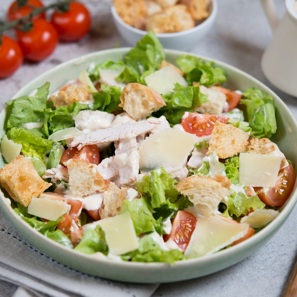 A vibrant Caesar salad with chicken, croutons, parmesan, and cherry tomatoes in a light green bowl.