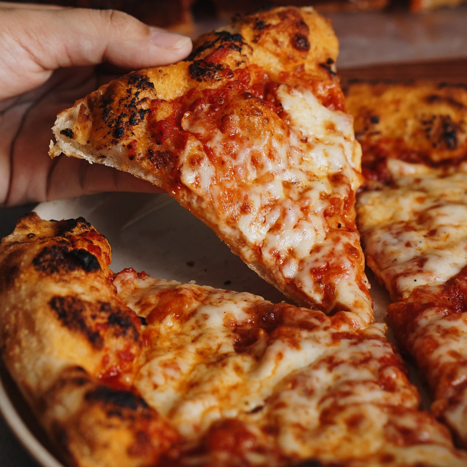 Hand lifting a slice of cheesy pizza with a charred crust from a whole pizza.