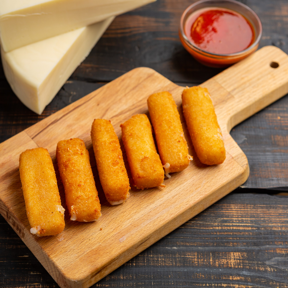 Six golden-brown mozzarella sticks on a wooden board, with a bowl of red sauce and cheese in the background.